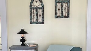 Een kamer met een blauwgroene fauteuil, een klein bijzettafeltje met een lamp, en twee decoratieve raamkozijnen aan de muur.