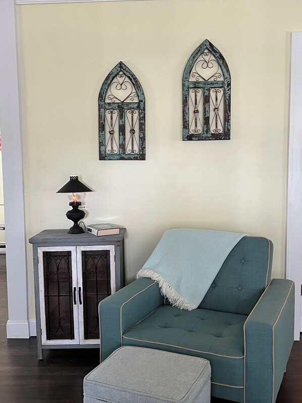 Ein Zimmer mit einem türkisfarbenen Sessel, einem kleinen Beistelltisch mit Lampe und zwei dekorativen Fensterrahmen an der Wand.