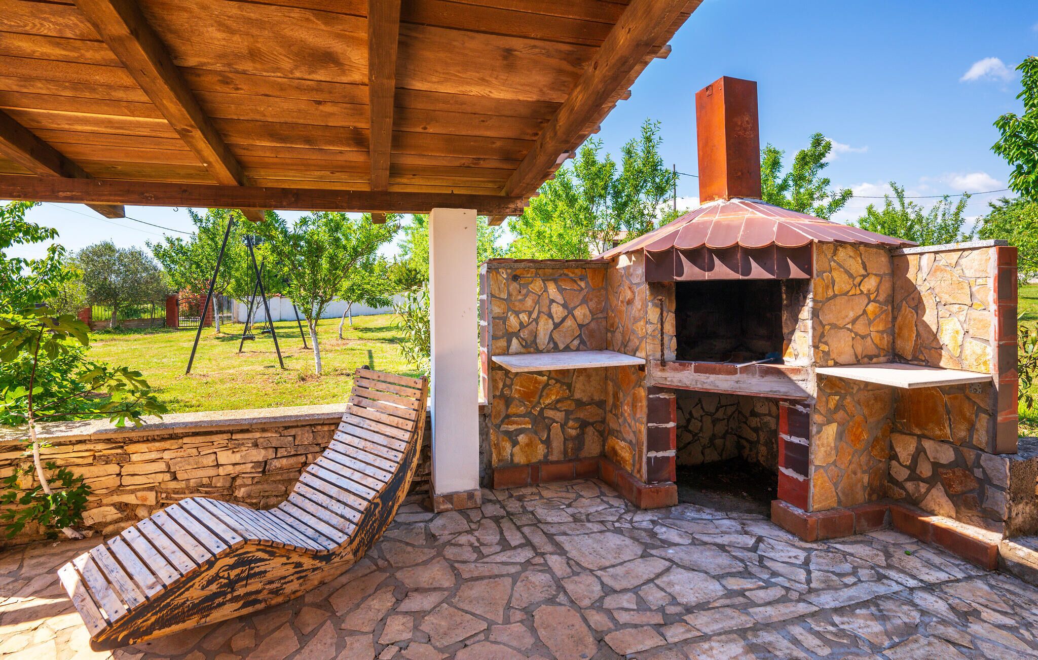 A stone-built outdoor kitchen with a chimney, a wooden bench, and a view of a grassy area with trees.