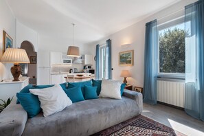 Una sala moderna con un sofá gris, cojines azules, un tapete estampado, una cocina al fondo y ventanas amplias con cortinas azules.