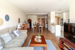 A modern living room with a sofa, coffee table, and TV. There is a dining area with a table and chairs. The kitchen is visible in the background. - Apartment 'Estrelinha Do Mar' with Shared Pool, Wi-Fi and Air Conditioning (Apúlia)