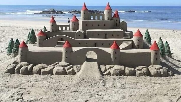 Un castillo de arena con múltiples torres y un puente, situado en una playa con el océano al fondo.