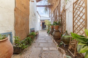 Un estrecho sendero de piedra bordeado por macetas con plantas y muebles rústicos, que conduce a la entrada de un edificio.