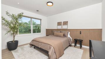 A modern bedroom with a large bed, a potted plant, a flat-screen TV, and a window with a view of greenery.