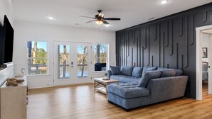 Un salon moderne avec un canapé gris, un plancher en bois, un ventilateur de plafond et de grandes fenêtres donnant sur la verdure.