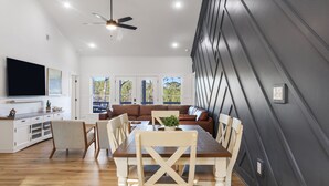 Un espace repas moderne avec une table et des chaises en bois, un ventilateur de plafond et une vue sur l’extérieur grâce à de grandes fenêtres.
