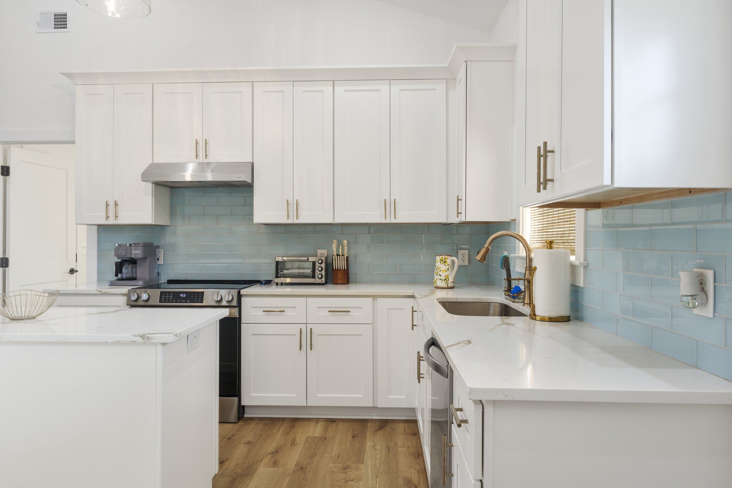 A modern kitchen with white cabinetry, a stainless steel range hood, a built-in oven, a microwave, and a sink with a gold faucet.