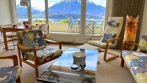 A cozy living room with a glass coffee table, floral-patterned chairs, and a view of a mountain and lake.