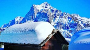Une cabane en bois dont le toit est recouvert de neige, entourée d’un paysage enneigé et avec une montagne en arrière-plan.