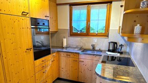 A compact kitchen with wooden cabinets, a built-in oven, a sink, and a window with a view of greenery. - Concierge, Scenic Views, Long Stay by SwissHut (Sigriswil)
