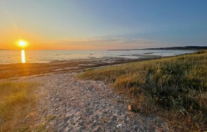 A beach scene with the sun setting over the water, a sandy path leading to the shore, and grassy areas. - 3 bedroom beautiful home in Hørve (Hørve)