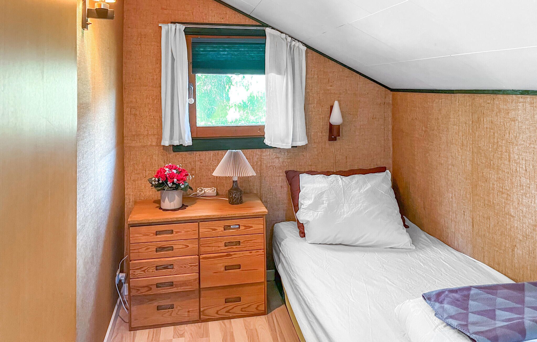 A small, cozy room with a bed, a wooden dresser with drawers, a window with curtains, and a lamp on the dresser.
