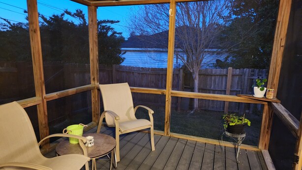 Un espacio cubierto con asientos al aire libre, con dos sillas y una mesita, que da a un patio trasero con terraza de madera y vista a un cuerpo de agua.