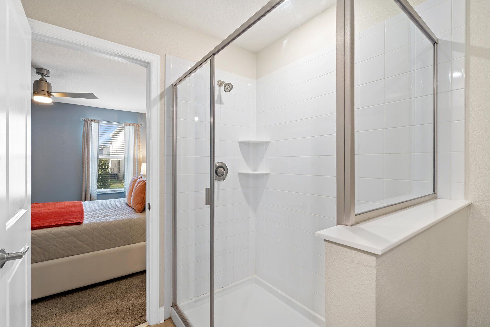 Une salle de bain moderne avec des carreaux blancs, une douche en verre et un lit visible dans la pièce adjacente, recouvert d’une couverture rouge.