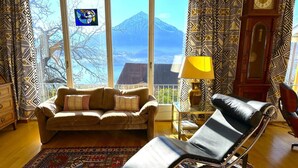 A living room with a sofa, a lounge chair, a patterned rug, a lamp, and a view of a mountain through large windows.