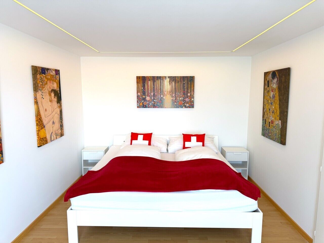 Une chambre moderne avec un lit blanc, des coussins rouges et deux tables de chevet. Trois tableaux ornent les murs.