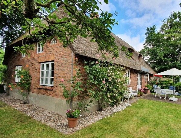 A brick house with a thatched roof, surrounded by a garden with flowering plants and a patio area with outdoor furniture.