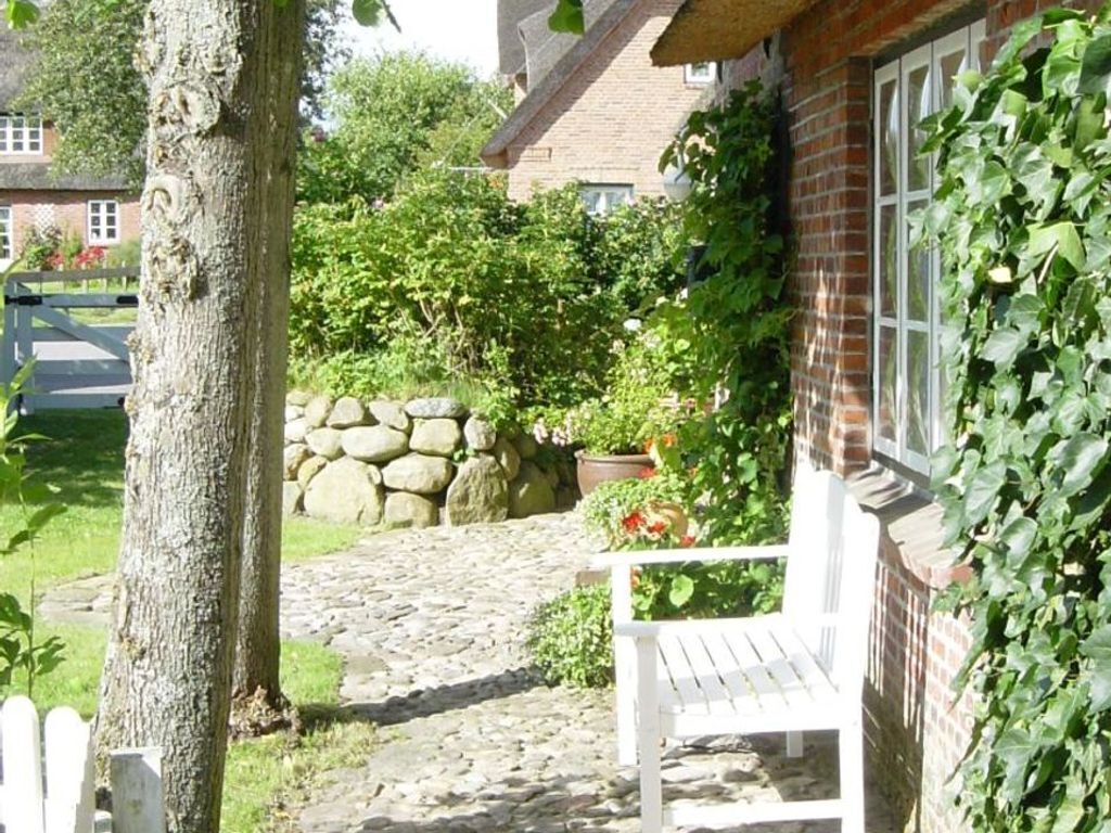 Een pad van stenen leidt naar een witte bank in een tuin, met een bakstenen gebouw, een stenen muur en begroeiing.