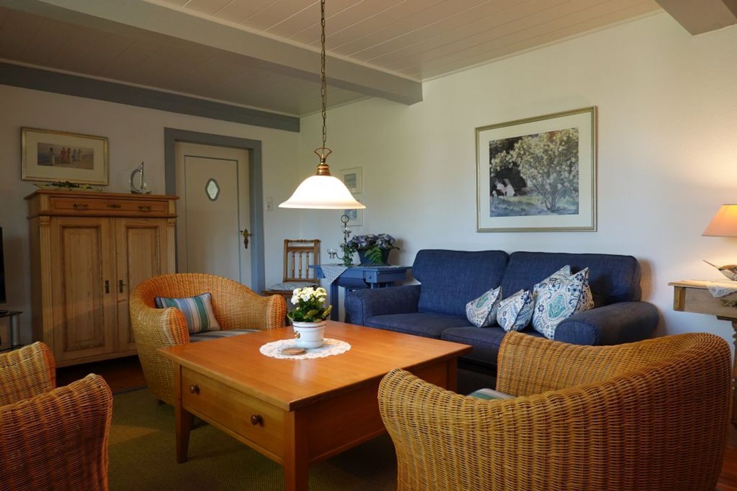 A cozy living room with a wooden dining table, wicker chairs, a blue sofa with patterned cushions, and a hanging lamp.