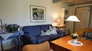 Un salón acogedor con un sofá azul, una mesa de comedor de madera, sillas de mimbre, una estantería y un cuadro en la pared.