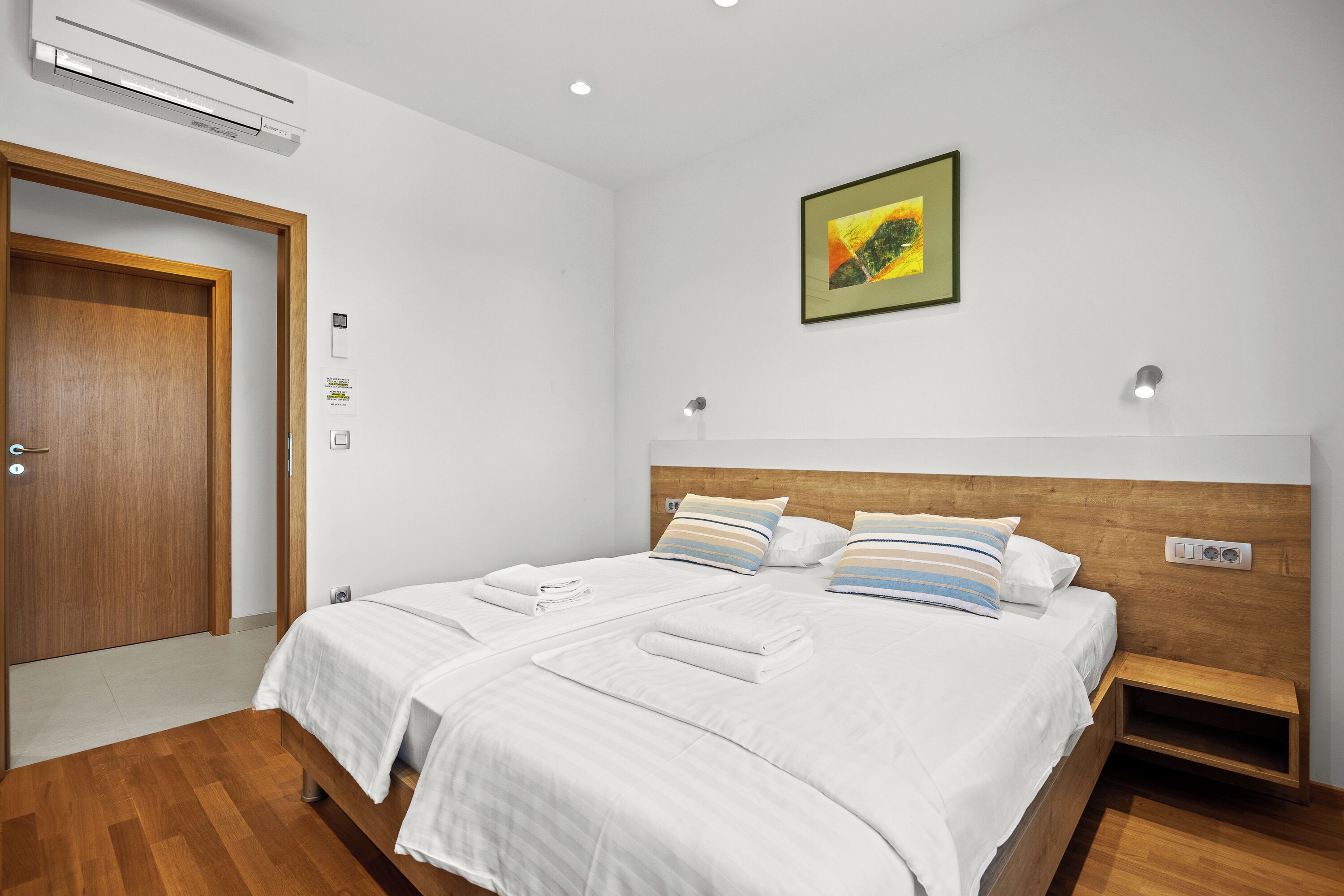 Une chambre d’hôtel avec un lit double, une tête de lit en bois, un climatiseur fixé au mur, un tableau encadré et une table de chevet.