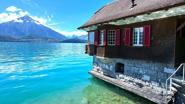 Ein Haus mit Naturstein-Fassade und roten Fensterläden, am Ufer eines klaren Sees mit Bergen im Hintergrund.