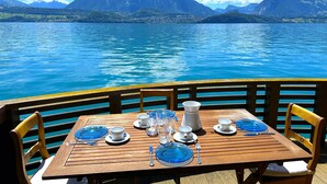 A dining table set with plates, glasses, and cutlery on a wooden deck overlooking a lake with mountains in the background. - Concierge, Boat House Scenic Views by SwissHut (Sigriswil)