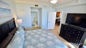 Ein Schlafzimmer mit Bett, Kommode, Fernseher und einem durchgehenden Türrahmen sichtbaren Badezimmer.