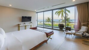 Een hotelkamer met een groot bed, een televisie, een zithoek met uitzicht op het strand en een balkon met gordijnen.