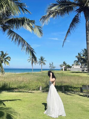 A woman in a white dress standing on a grassy area with palm trees and a view of the ocean. - The Five Villas Danang (Da Nang)