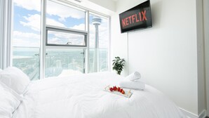 A hotel room with a large bed, a view of the cityscape through floor-to-ceiling windows, a wall-mounted TV displaying the Netflix logo, and a tray of fruit on the bed.