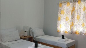 A room with two single beds, a bedside table with a lamp, a window with yellow floral curtains, and an air conditioning unit on the wall. - BC Lodge (Puerto Princesa)