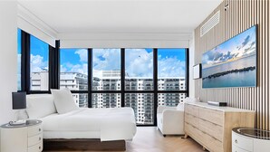 Ein modernes Hotelzimmer mit einem großen Bett, einem Fernseher, einer Kommode und Blick auf die Stadt durch große Fenster.