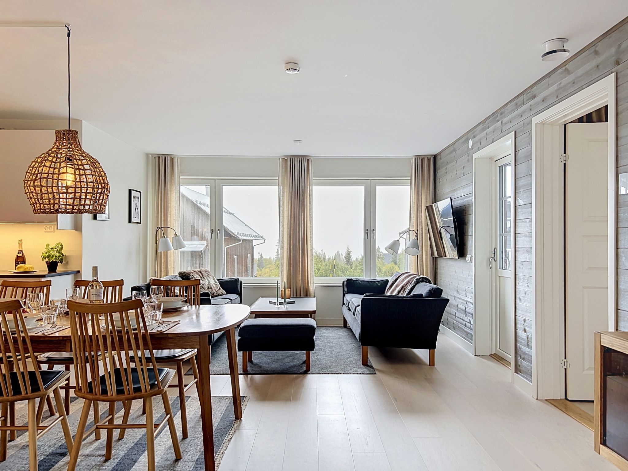 A modern living room with a dining area, large windows, and a flat-screen TV mounted on the wall.