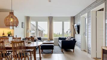 A modern living room with a dining area, large windows, and a flat-screen TV mounted on the wall.