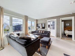 Una sala de estar moderna con sofá, reposapiés y mesa de centro, ventanas amplias y un televisor de pantalla plana montado en la pared.