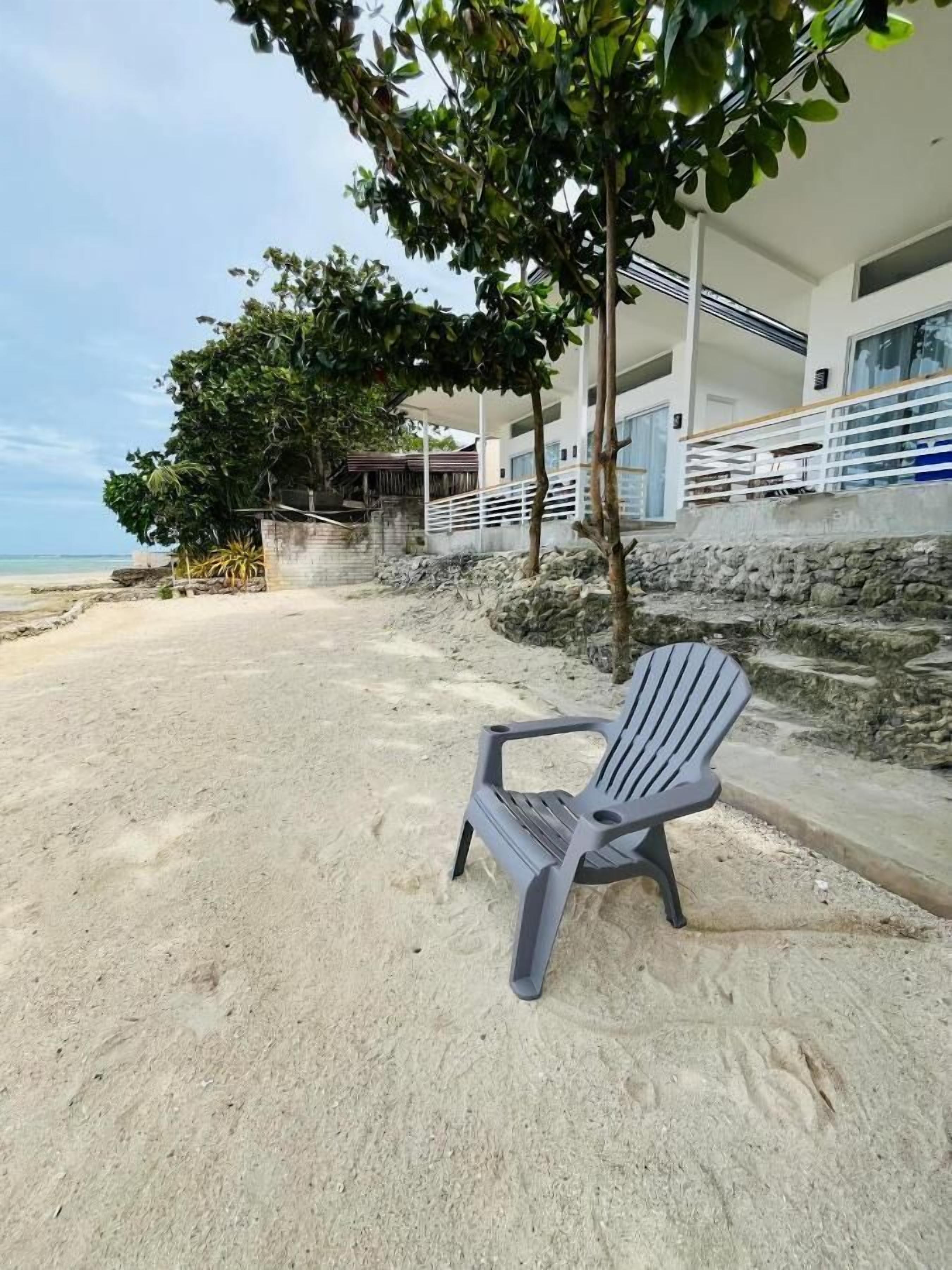 A beachside hotel with a sandy beach, a single grey plastic chair, and a building with a balcony overlooking the sea.