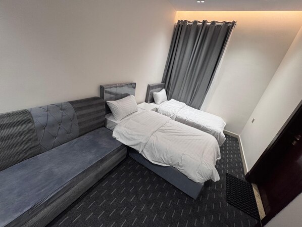 Una habitación de hotel moderna con un sofá gris, dos camas individuales, una mesita pequeña y una cortina.