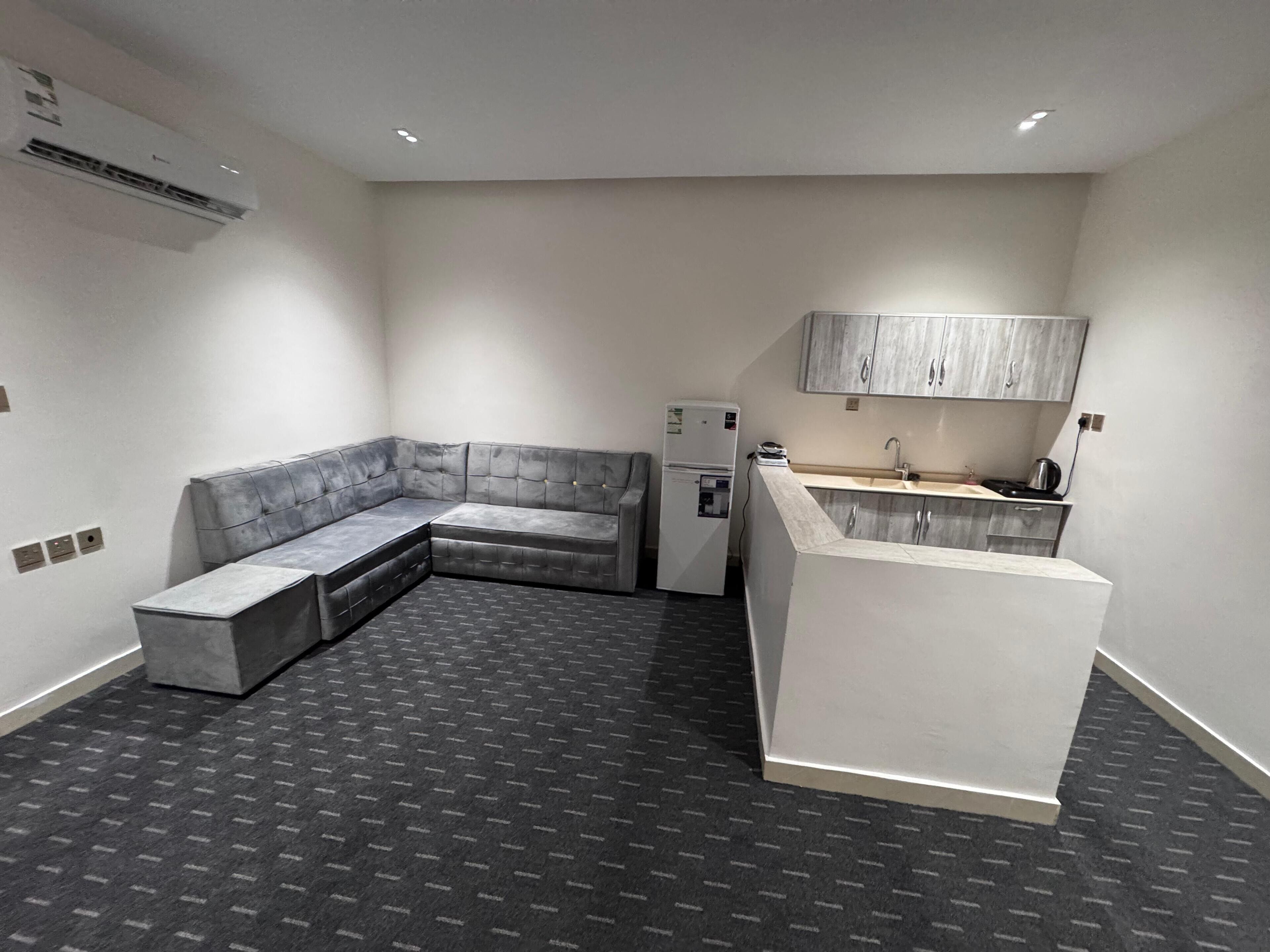 Ein modernes, kompaktes Wohnzimmer mit grauem Ecksofa, einer kleinen Küchenzeile und einer eingebauten Sitzbank.