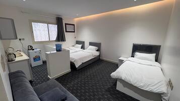 A hotel room with two single beds, a small kitchenette, a sofa, and a window with curtains.