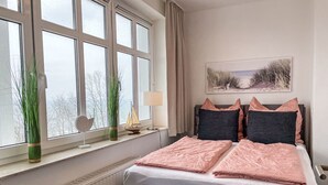 Un dormitorio con un ventanal grande, un radiador, una mesita de noche con una lámpara y una cama con ropa de cama rosa y gris.