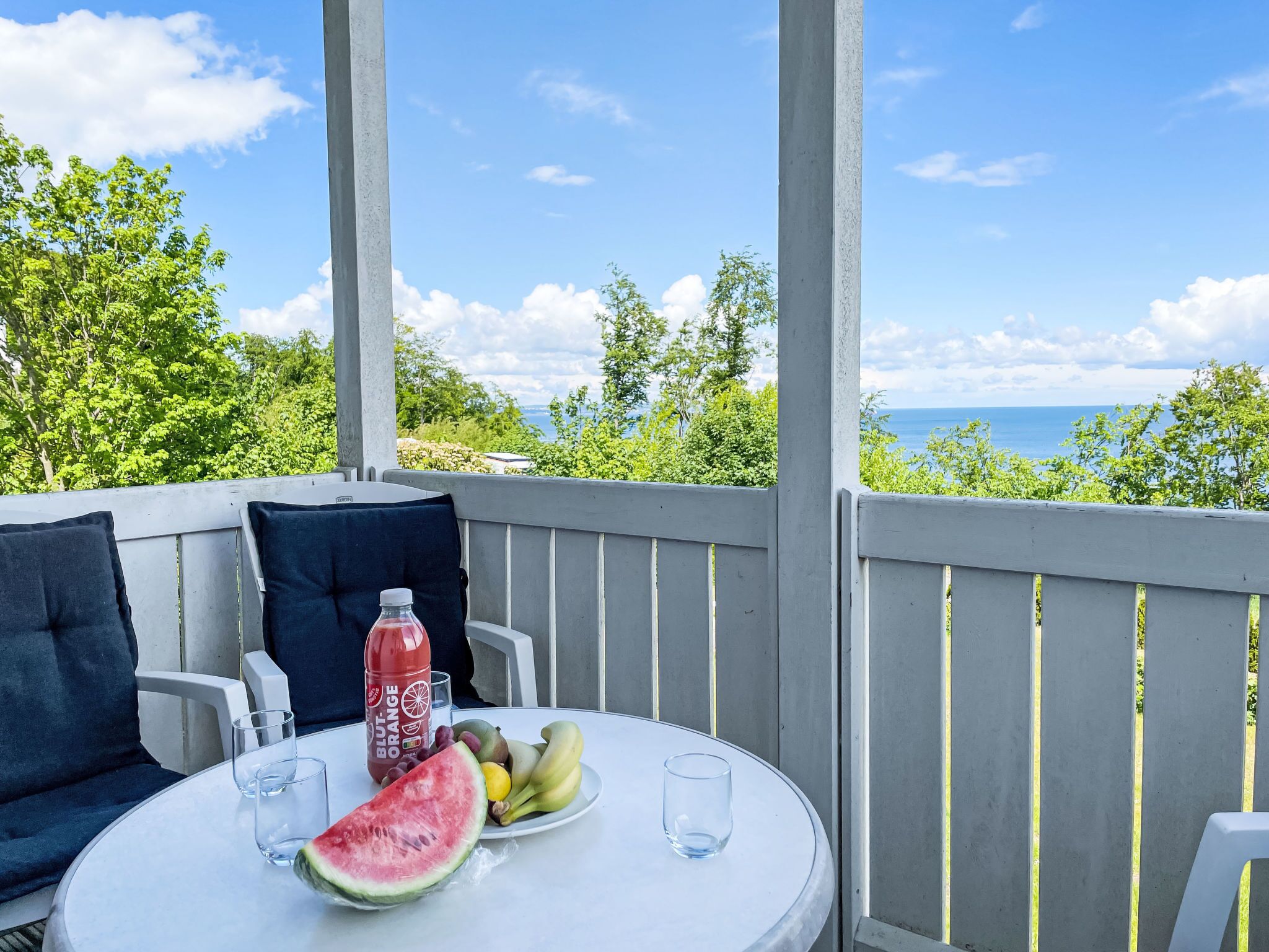 Ein Tisch mit einer Wassermelone, Obst und einer Flasche auf einem Balkon mit Blick aufs Meer.