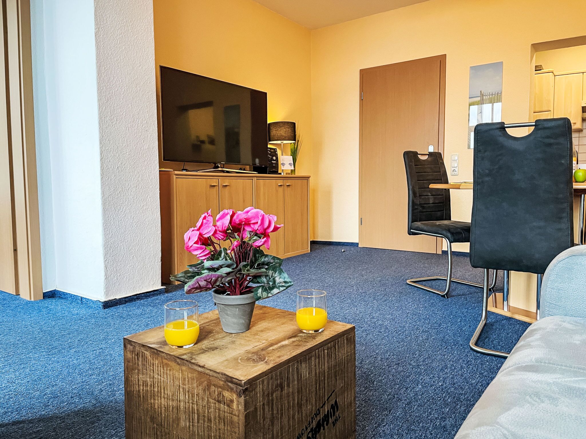 A hotel room with a wooden coffee table, two glasses of orange juice, a potted plant with pink flowers, a black chair, and a TV on a wooden cabinet.