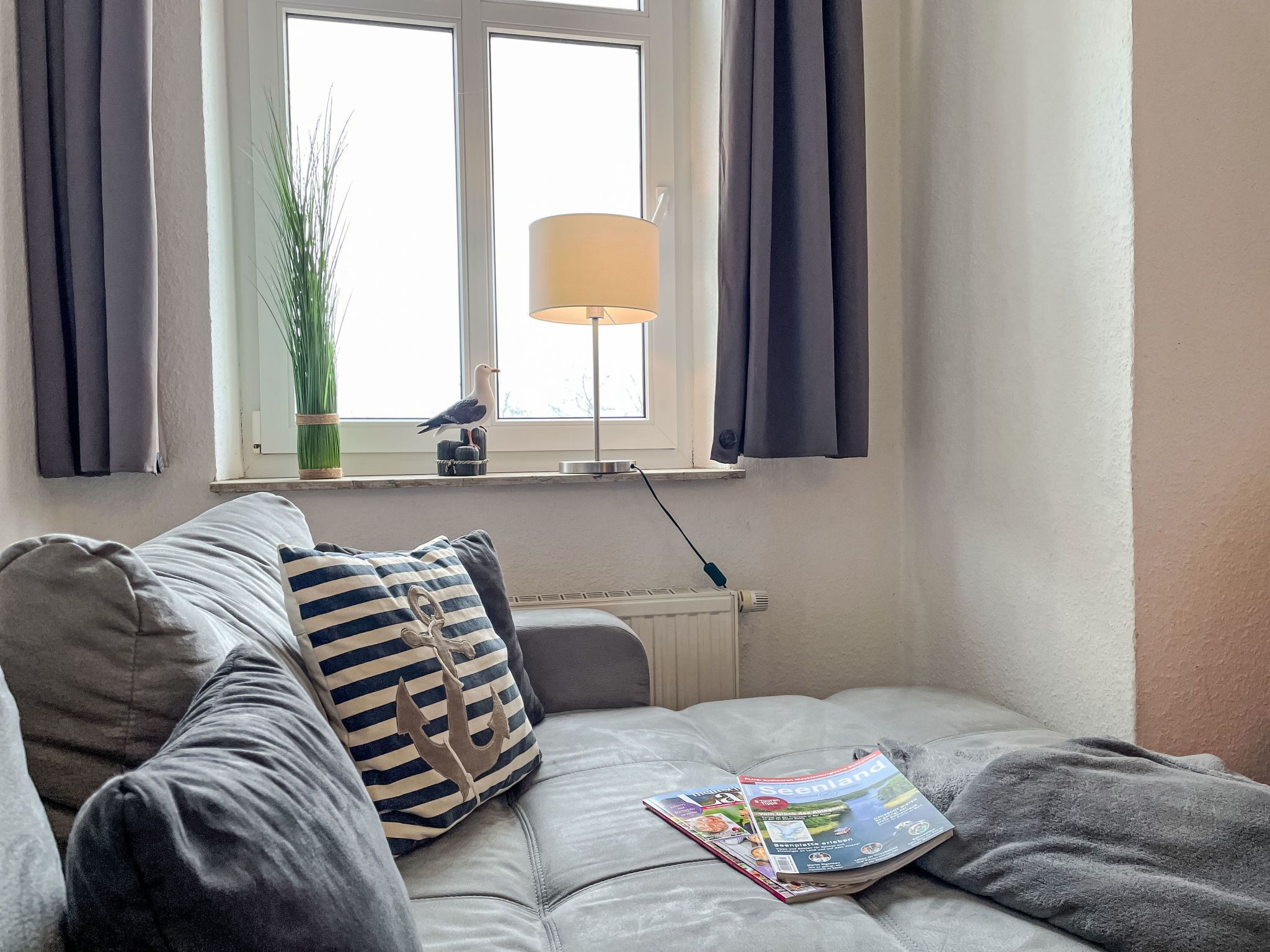 Un salon chaleureux avec un canapé gris, un coussin à rayures et un magazine posé dessus. On y trouve une fenêtre avec des rideaux, une lampe et une plante sur le rebord de la fenêtre.