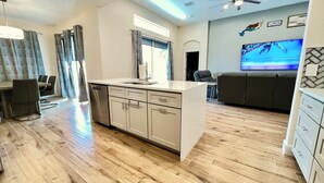A modern kitchen with a central island, a dining area with chairs, a sofa, and a TV displaying a beach scene. - Highlands Reserve Villa with screened-in patio & pool. Peaceful Golf Community. (Davenport)