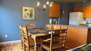 A dining area with a wooden table and chairs, a chandelier, and a kitchen with wooden cabinets and a refrigerator.