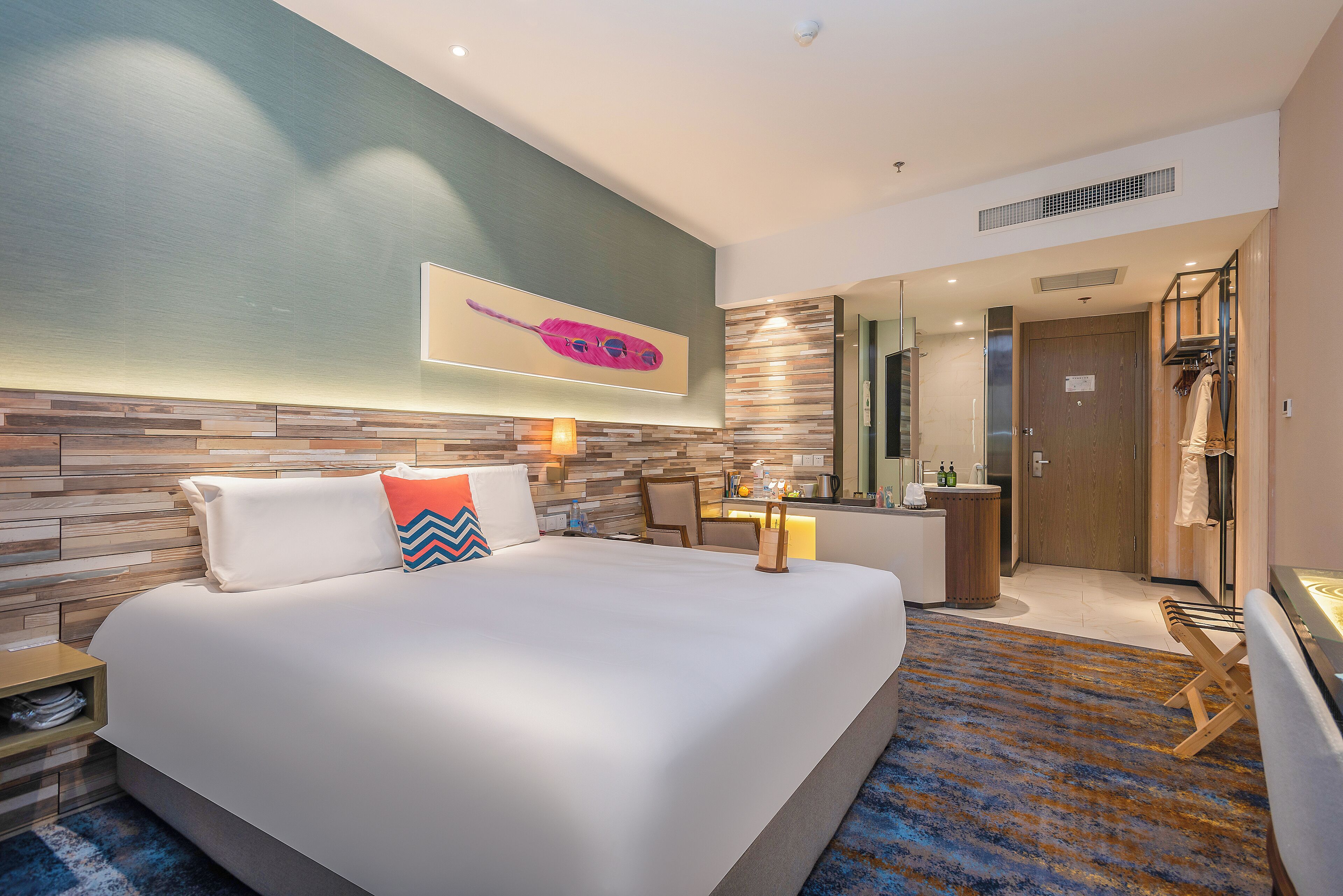 A modern hotel room with a large bed, a wooden headboard, a nightstand, a desk with a chair, a bathroom with a mirror, and a shower area.