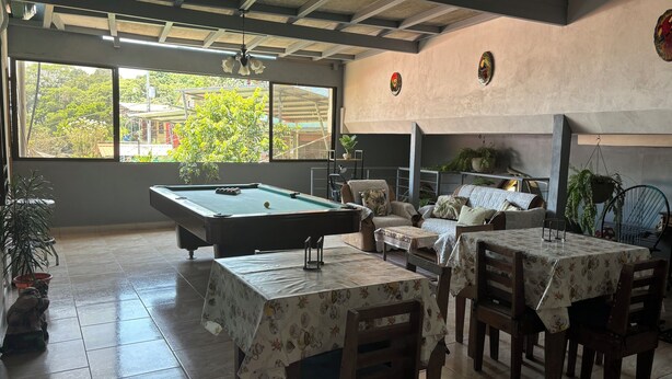 Une table de billard dans une pièce dotée de grandes fenêtres, d’un canapé et de plusieurs tables recouvertes de nappes à motifs floraux.