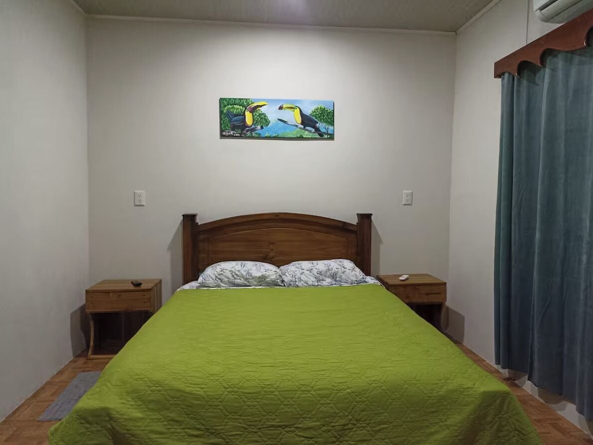 Ein Schlafzimmer mit einem hölzernen Bett, einer grünen Bettdecke, zwei Nachttischen, einem Wandbild von Tukane und einem Fenster mit Vorhängen.