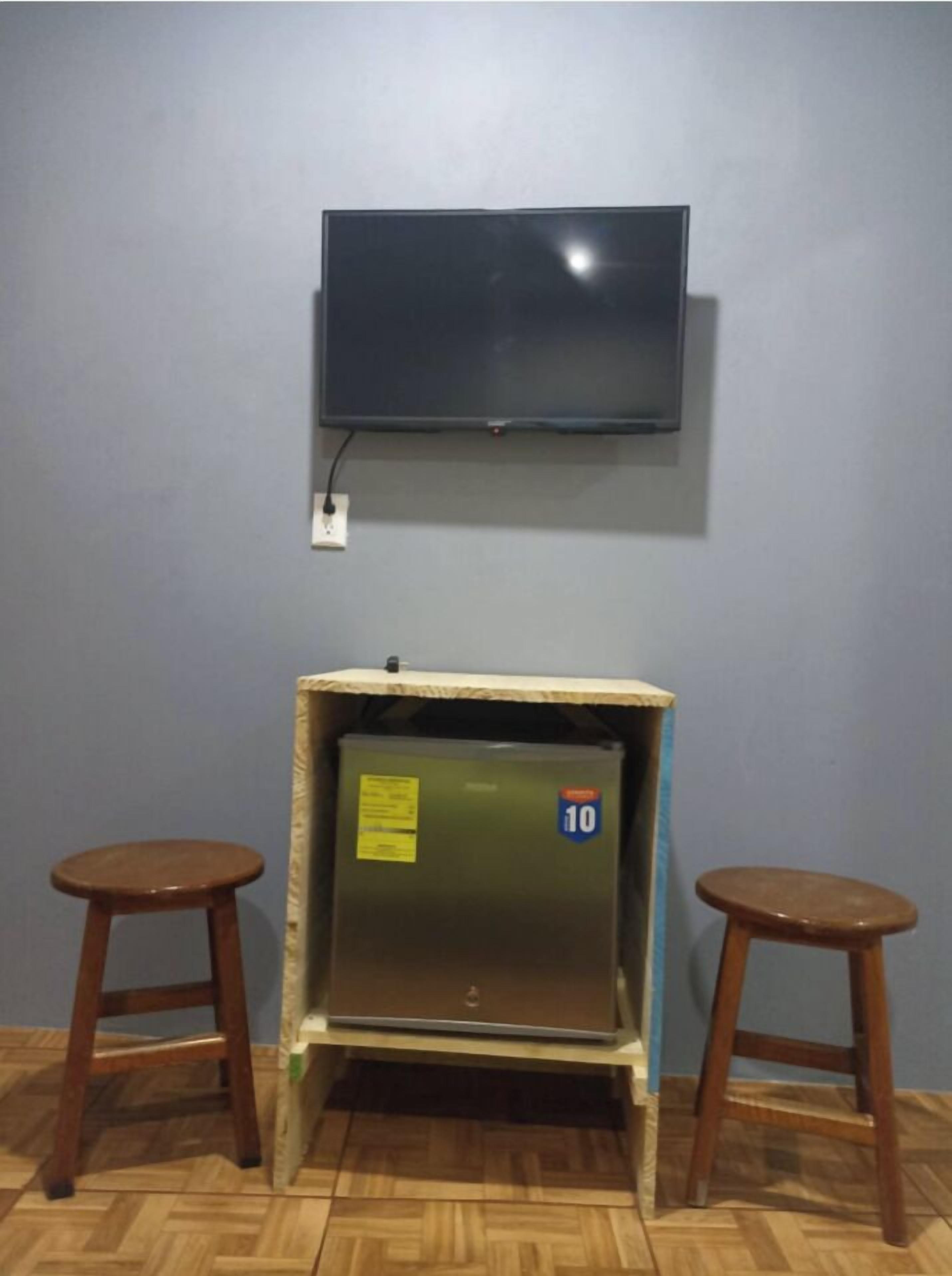 Een kamer met een wandgemonteerde flatscreen-tv, een ingebouwde minibar en twee houten krukken.